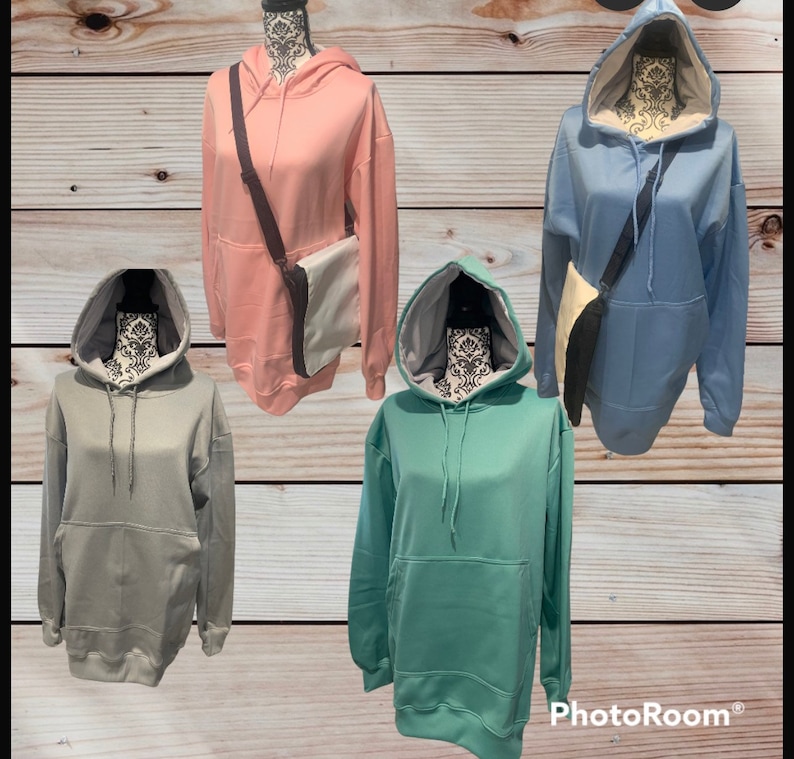 Op de afbeelding: Vier hoodies in verschillende kleuren: roze, lichtblauw, grijs en teal. Elke hoodie heeft een zak met een kangoeroe-stijl en een capuchon met een trekkoord. De hoodies worden getoond op mannequins met een bruine houten achtergrond.