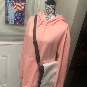 Op de afbeelding: Een roze hoodie met een zwarte band en een witte schoudertas. De tas heeft een zwarte rits en een zwarte bies.
