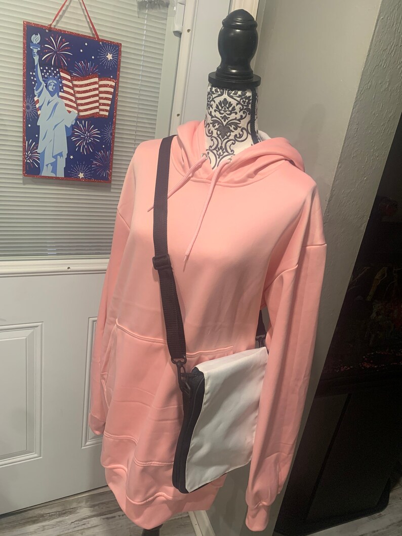 Op de afbeelding: Een roze hoodie met een zwarte band en een witte schoudertas. De tas heeft een zwarte rits en een zwarte bies.