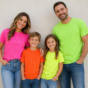 May include: A group of four people wearing short-sleeved crew neck t-shirts in bright neon colors including hot pink, orange, and lime green, paired with blue denim jeans. The shirts are solid-colored with a simple, standard fit and design.