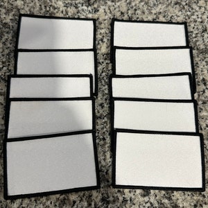 May include: A set of ten blank white iron-on patches with black borders. The patches are rectangular and have a smooth surface.