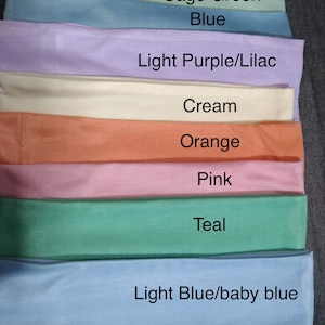 May include: A stack of eight fabric headbands in different colors, including sage green, blue, light purple, cream, orange, pink, teal, and light blue. The colors are labeled below each headband.