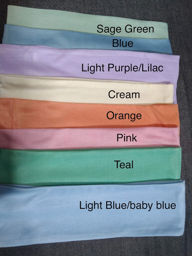 May include: A stack of eight fabric headbands in different colors, including sage green, blue, light purple, cream, orange, pink, teal, and light blue. The colors are labeled below each headband.