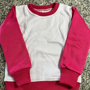 May include: A white and pink sweatshirt with a crew neck. The sleeves and bottom hem are a vibrant pink, while the body is white. The neckline is trimmed with a thin red band.