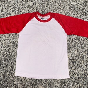 Può includere: Una maglietta da baseball bianca e rossa a maniche corte. La maglietta ha un corpo bianco e maniche rosse.