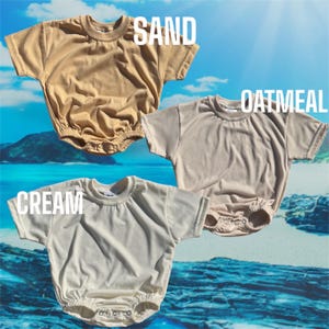 May include: Three baby onesies in different colors: sand, oatmeal, and cream. The onesies are made of a soft, comfortable fabric and have a classic, simple design.