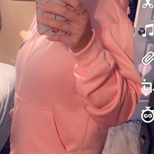 Op de afbeelding: Een roze hoodie met een grote zak aan de voorkant. De hoodie wordt gedragen door een persoon, maar het gezicht van de persoon is bedekt door een telefoon.