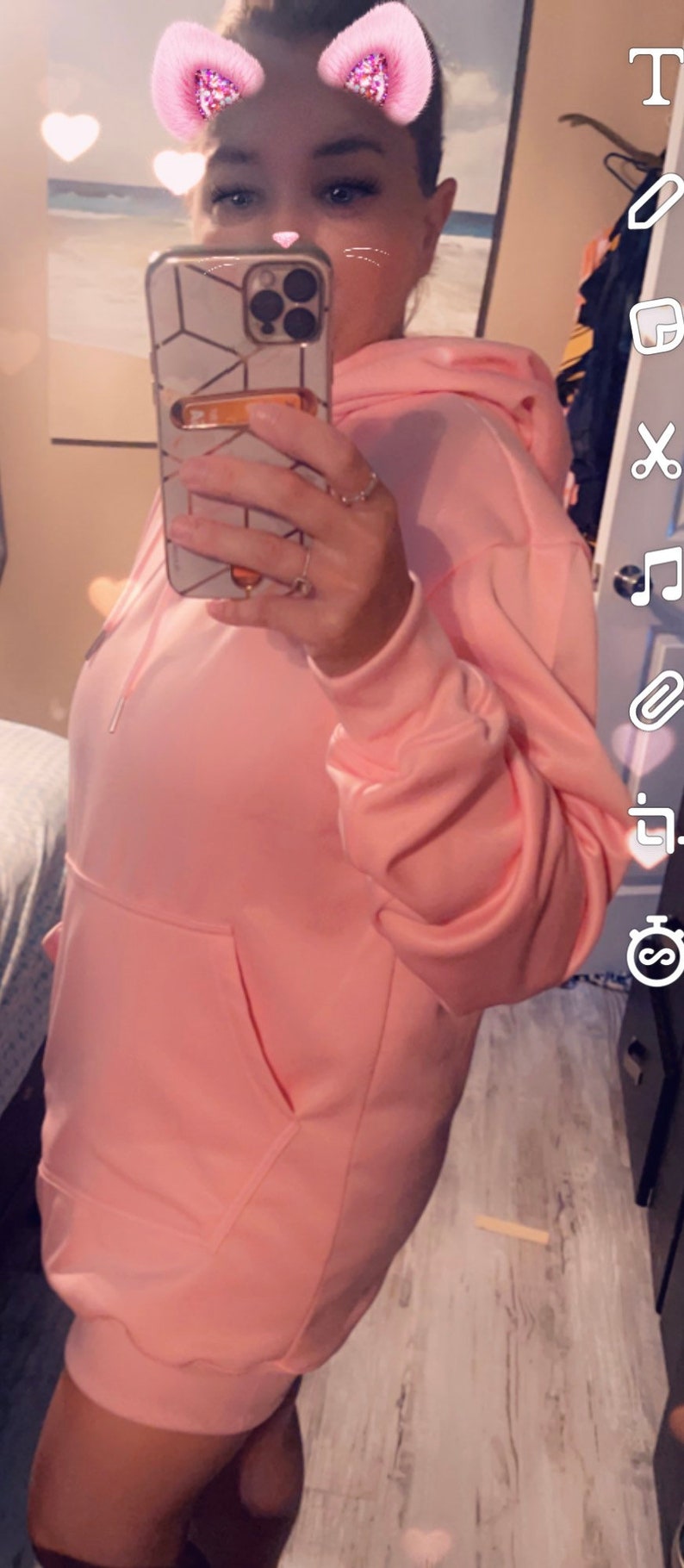Op de afbeelding: Een roze hoodie met een grote zak aan de voorkant. De hoodie wordt gedragen door een persoon, maar het gezicht van de persoon is bedekt door een telefoon.