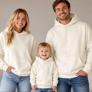 May include: Three matching cream-colored hooded sweatshirts with long sleeves, front kangaroo pockets, and ribbed cuffs. Each sweatshirt features a hood and a plain, solid-colored design, worn with blue denim jeans by the individuals in the image.