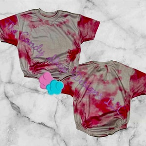 May include: Two tie-dye shirts in a light beige color with a red and pink tie-dye pattern. The shirts have short sleeves and a round neckline.