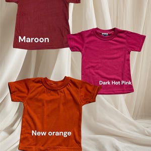 May include: Three plain t-shirts in maroon, dark hot pink, and new orange. The shirts are laid out on a white fabric background.