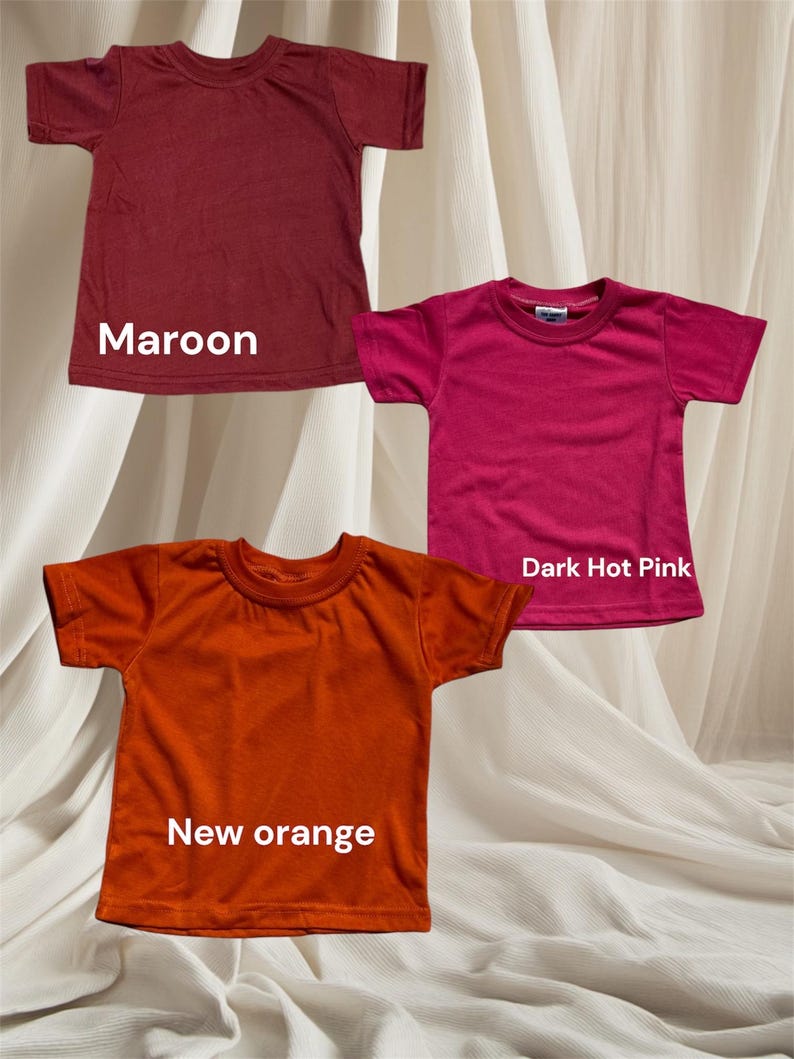 May include: Three plain t-shirts in maroon, dark hot pink, and new orange. The shirts are laid out on a white fabric background.