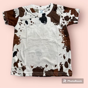 May include: White short-sleeved t-shirt with a brown cowhide pattern. The shirt has a crew neck and a tag that reads "THE COMFY SHOP DESIGNS LLC". The cow print design covers the front and back of the shirt.