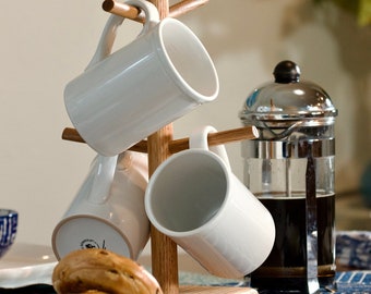 May include: A wooden mug rack with four white ceramic mugs hanging from it. The rack is on a table with a French press and two bagels.