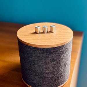 May include: A cylindrical speaker with a wooden top and base, and a dark gray fabric-like exterior. Three silver knobs are centered on the top. The speaker sits on a wooden surface against a blue wall.