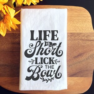 May include: White kitchen towel with black text that reads "Life is Short Lick the Bowl".