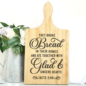May include: Wooden cutting board with a brown wood grain finish. The board has a handle and a quote engraved on it: "They broke bread in their homes and ate together with glad and sincere hearts. Acts 2:46."