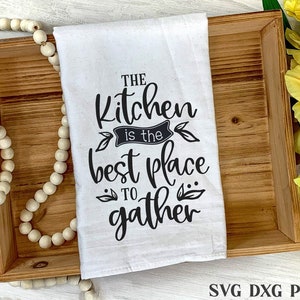 May include: A white kitchen towel with black text that reads "The Kitchen is the best place to gather". The towel is displayed on a wooden tray with a string of white beads.