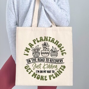 May include: A white tote bag with a green and black design that says "I'm a plantanolic on the road to recovery, just kidding, I'm on my way to get more plants".