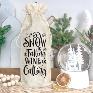 May include: A burlap wine bag with the text "Snow is Falling Wine is Calling" in black lettering. The bag is sitting on a table with a snow globe and other winter decorations.