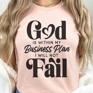 May include: A pink t-shirt with the text "God is within my Business Plan I will not Fail" in black lettering.