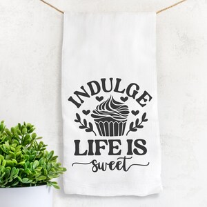 May include: A white kitchen towel with black text that reads "Indulge Life is sweet" with a cupcake graphic in the center.
