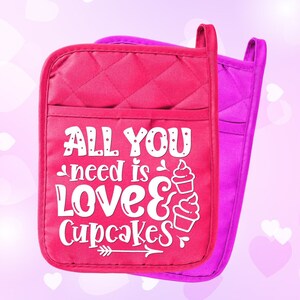 May include: A pair of pink oven mitts with a white quilted design. The mitts have a white graphic that says "All you need is love & cupcakes" with a cupcake graphic and an arrow.