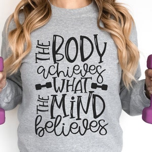 Puede incluir: Una sudadera gris con el texto "The body achieves what the mind believes" en letras negras. El texto est&aacute; escrito a mano. La sudadera est&aacute; siendo sostenida por una persona que sostiene pesas rosadas.