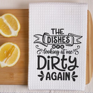 May include: A white waffle-weave kitchen towel with the text "THE DISHES are looking at me Dirty AGAIN" in black. Two lemon halves sit on a wooden cutting board, with a knife to the right.