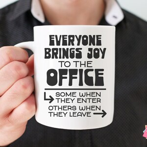 Puede incluir: Taza de cer&aacute;mica blanca con texto negro que dice "Todo el mundo trae alegr&iacute;a a la oficina. Algunos cuando entran, otros cuando se van." La taza est&aacute; siendo sostenida por la mano de una persona.