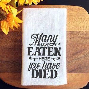 May include: White kitchen towel with black text that reads "Many have eaten here few have died".