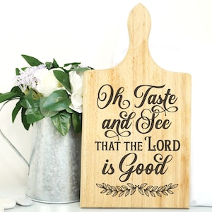 May include: Wooden cutting board with the text "Oh Taste and See That The Lord is Good" in a decorative font. The board has a handle and is decorated with a leafy design.
