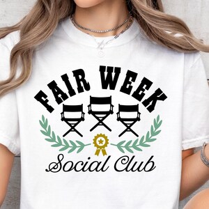 May include: White t-shirt with black text that reads "Fair Week Social Club". The text is surrounded by a wreath of green leaves and three black director's chairs.