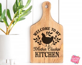 Könnte beinhalten: Holzschneidebrett mit einer schwarzen Silhouette eines Huhns und dem Text "Willkommen in meiner Mother Cluckin' Küche".