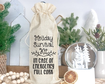 Könnte beinhalten: Ein Weinsack aus Jute mit dem Text "Holiday Survival Kit. In Case of Emergency Pull Cork". Der Sack liegt auf einem Tisch mit einer Schneekugel und anderen Weihnachtsdekorationen.