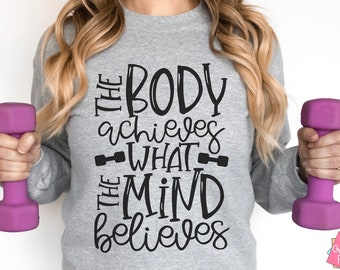 May include: A gray sweatshirt with the text "The body achieves what the mind believes" in black lettering. The text is in a handwritten style. The sweatshirt is being held by a person holding pink dumbbells.