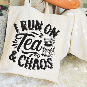 May include: A white tote bag with black text that reads "I run on tea & chaos". The text is in a stylized font and there are three stacked teacups with a black and white pattern.