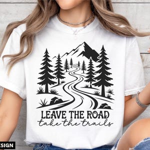May include: White t-shirt featuring a black graphic of a mountain scene with a winding road and pine trees. The text "LEAVE THE ROAD take the trails" is below the graphic. Digital design.