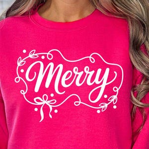 May include: A bright pink sweatshirt with the word "Merry" in white script, framed by a decorative border of leaves and bows. The sweatshirt is a festive holiday apparel item.