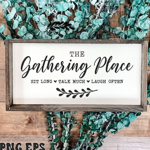 Op de afbeelding: Een wit houten bord met de tekst "THE Gathering Place SIT LONG TALK MUCH LAUGH OFTEN" in zwarte letters. Het bord is ingelijst in een donker houten frame en ligt op een houten oppervlak met groen blad.