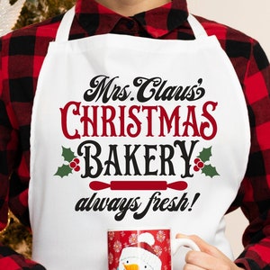 K&ouml;nnte beinhalten: Eine wei&szlig;e Sch&uuml;rze mit dem Text "Mrs. Claus's Christmas Bakery always fresh!" in schwarzer und roter Schrift. Die Sch&uuml;rze hat eine rote Nudelholzgrafik und gr&uuml;nen Stechpalmen mit roten Beeren. Eine Person tr&auml;gt die Sch&uuml;rze und h&auml;lt eine wei&szlig;e Tasse mit einem Schneemann-Design.