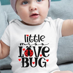 Puede incluir: Un body blanco con el texto "little miss love bug" en negro con un gráfico de mariquita roja. El body lo lleva una niña con una diadema morada.