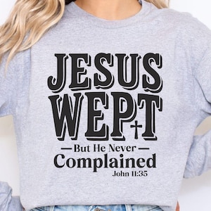 May include: A light gray long-sleeve sweatshirt with the text "JESUS WEPT" in a bold, black font, with a cross symbol. Below the text is "But He Never Complained" and "John 11:35". The words are in a stylized font.