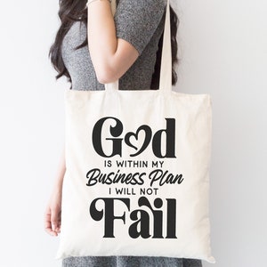 May include: A white tote bag with black text that reads "God is within my Business Plan I will not Fail". The text is in a stylized font with a heart in the middle of the word "God".