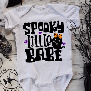 May include: White baby onesie with black text that reads "Spooky Little Babe" and a black ghost with a purple bow and a smiling face.