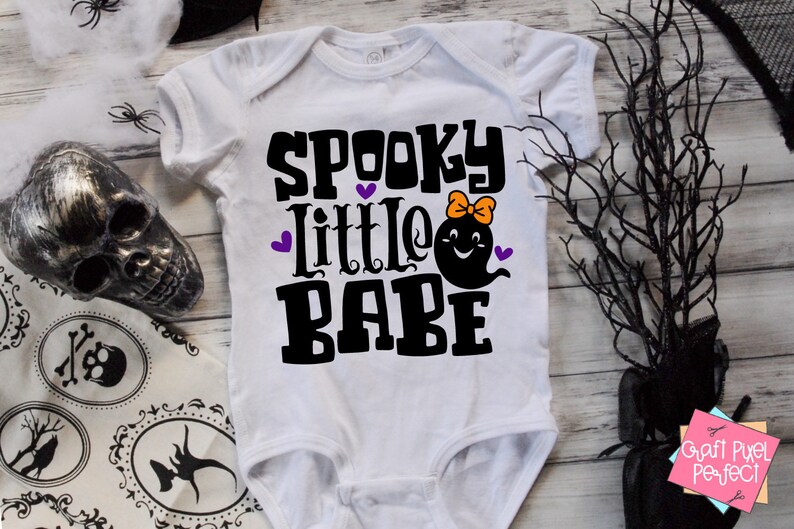 May include: White baby onesie with black text that reads "Spooky Little Babe" and a black ghost with a purple bow and a smiling face.