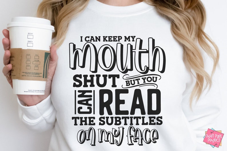 K&ouml;nnte beinhalten: Wei&szlig;es Sweatshirt mit dem Text "I can keep my mouth shut but you can read the subtitles on my face" in Schwarz. Eine Person h&auml;lt eine wei&szlig;e Kaffeetasse mit brauner H&uuml;lle.