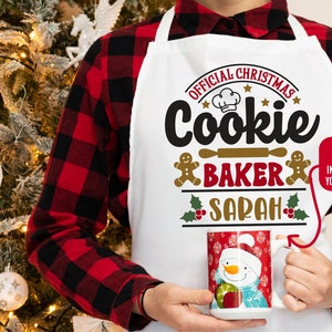 May include: White Christmas-themed apron with the text "Official Christmas Cookie Baker Sarah" in red, gold, and black lettering. A gingerbread man graphic is on either side of the text. A red mug with a snowman graphic is in the lower right corner.