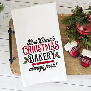 K&ouml;nnte beinhalten: Wei&szlig;es K&uuml;chentuch mit rotem und schwarzem Text, der "Mrs. Claus's Christmas Bakery always fresh!" lautet, mit einem roten Nudelholz und Stechpalmenbl&auml;ttern. Das Tuch liegt auf einem Holztablett mit rot-wei&szlig; karierten F&auml;ustlingen und einer roten Glocke mit gr&uuml;nen Kiefern&auml;sten.