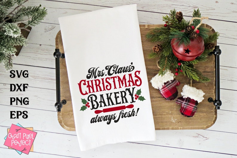 K&ouml;nnte beinhalten: Wei&szlig;es K&uuml;chentuch mit rotem und schwarzem Text, der "Mrs. Claus's Christmas Bakery always fresh!" lautet, mit einem roten Nudelholz und Stechpalmenbl&auml;ttern. Das Tuch liegt auf einem Holztablett mit rot-wei&szlig; karierten F&auml;ustlingen und einer roten Glocke mit gr&uuml;nen Kiefern&auml;sten.